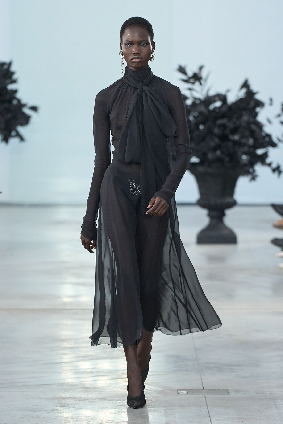 Model walking a sheer black gown with a large neck-tie knot on a runway.
