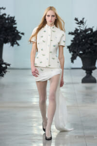 Model walks the runway in a cream floral short-sleeve jacket and draped skirt, black heels upcoming pose.