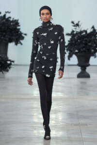 Model on a runway wearing a black turtleneck tunic covered in silver floral embellishments, with a black headwrap and matching ankle boots.