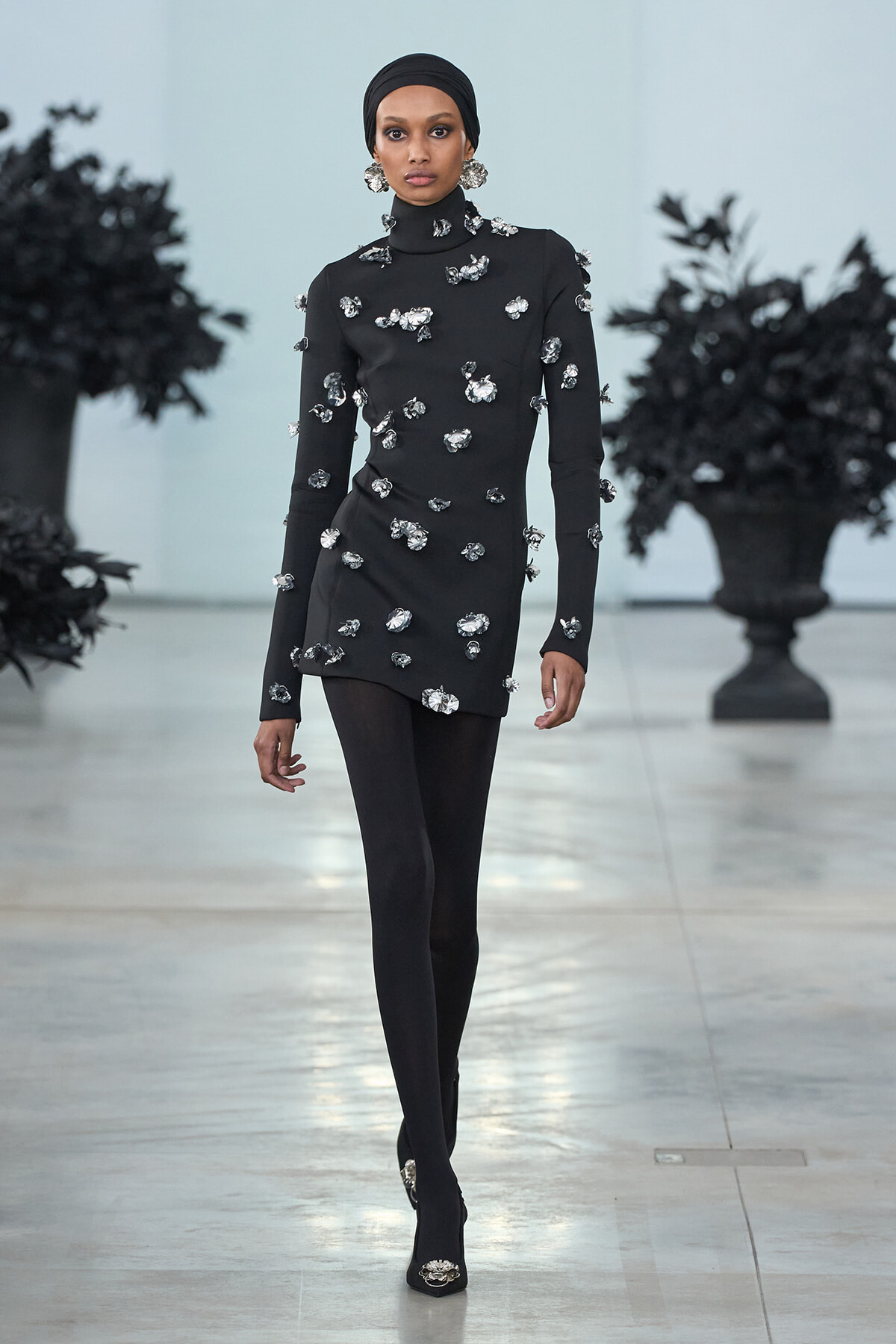 Model on a runway wearing a black turtleneck tunic covered in silver floral embellishments, with a black headwrap and matching ankle boots.