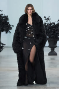 Model on a runway wearing a long black fur coat over a sheer black dress adorned with large silver crystals and a high slit.