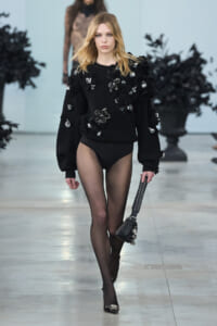 Model walking runway in black embellished sweater with floral appliqués, sheer tights and high heels, carrying a studded black handbag.