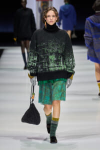 Model walks the runway in a black turtleneck oversized sweater with green speckle pattern and emerald green textured shorts, carrying a black beaded bag.