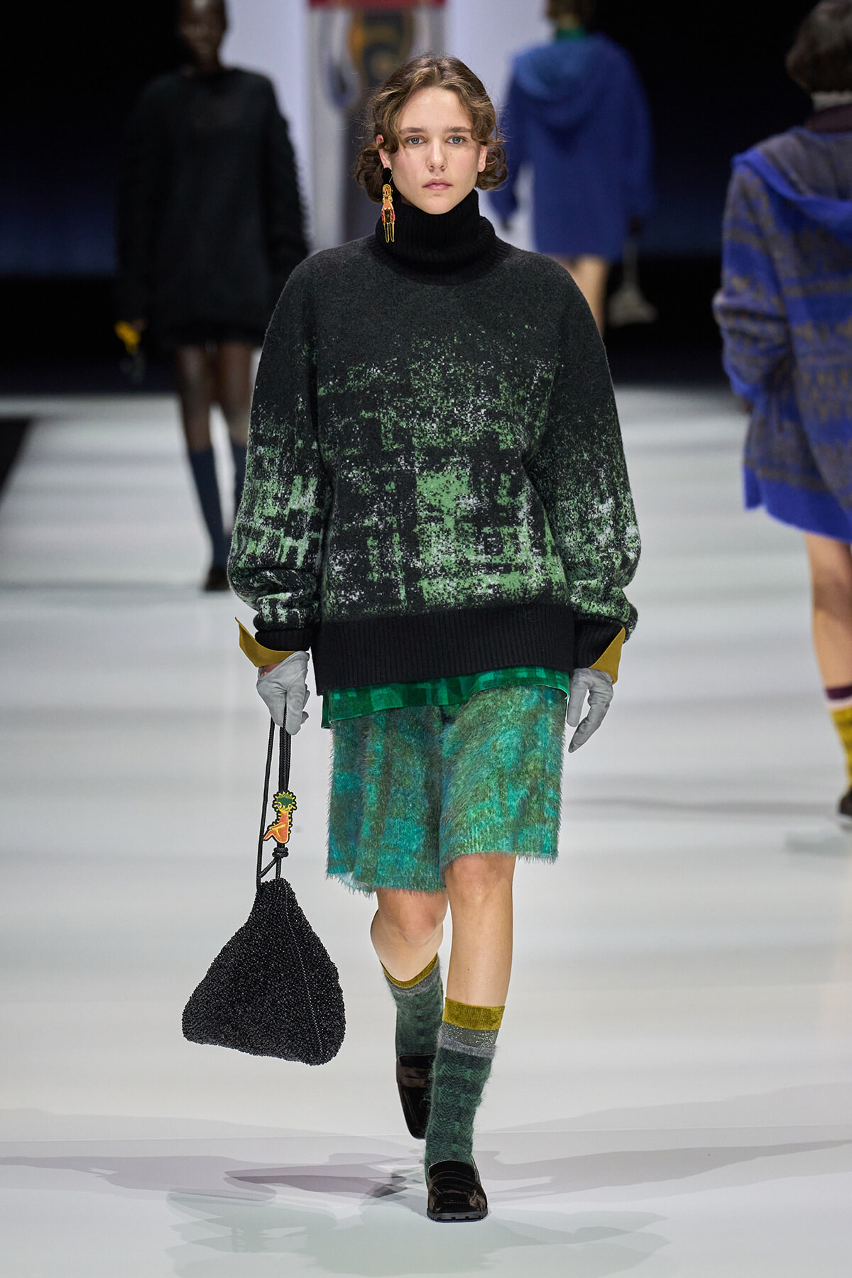 Model walks the runway in a black turtleneck oversized sweater with green speckle pattern and emerald green textured shorts, carrying a black beaded bag.