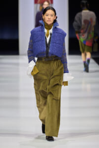 Female model walks a fashion runway in a blue textured cropped cardigan over a mustard turtleneck and white shirt, paired with olive cargo trousers and a brown belt.