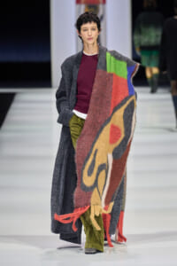 Model on a runway wearing a long gray cardigan over a burgundy top, with a large multicolored scarf draped over her shoulders.