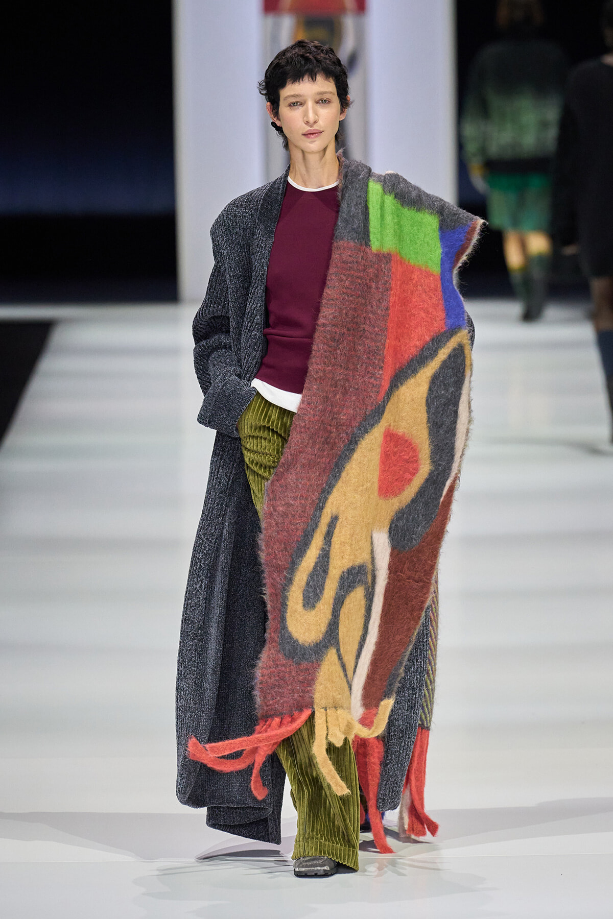 Model on a runway wearing a long gray cardigan over a burgundy top, with a large multicolored scarf draped over her shoulders.