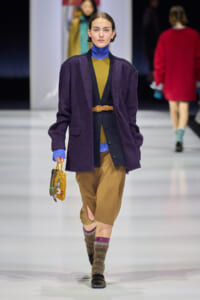 Model walking a fashion runway in a layered outfit: purple coat over mustard and blue knitwear, tan culottes, brown belt, beaded handbag.