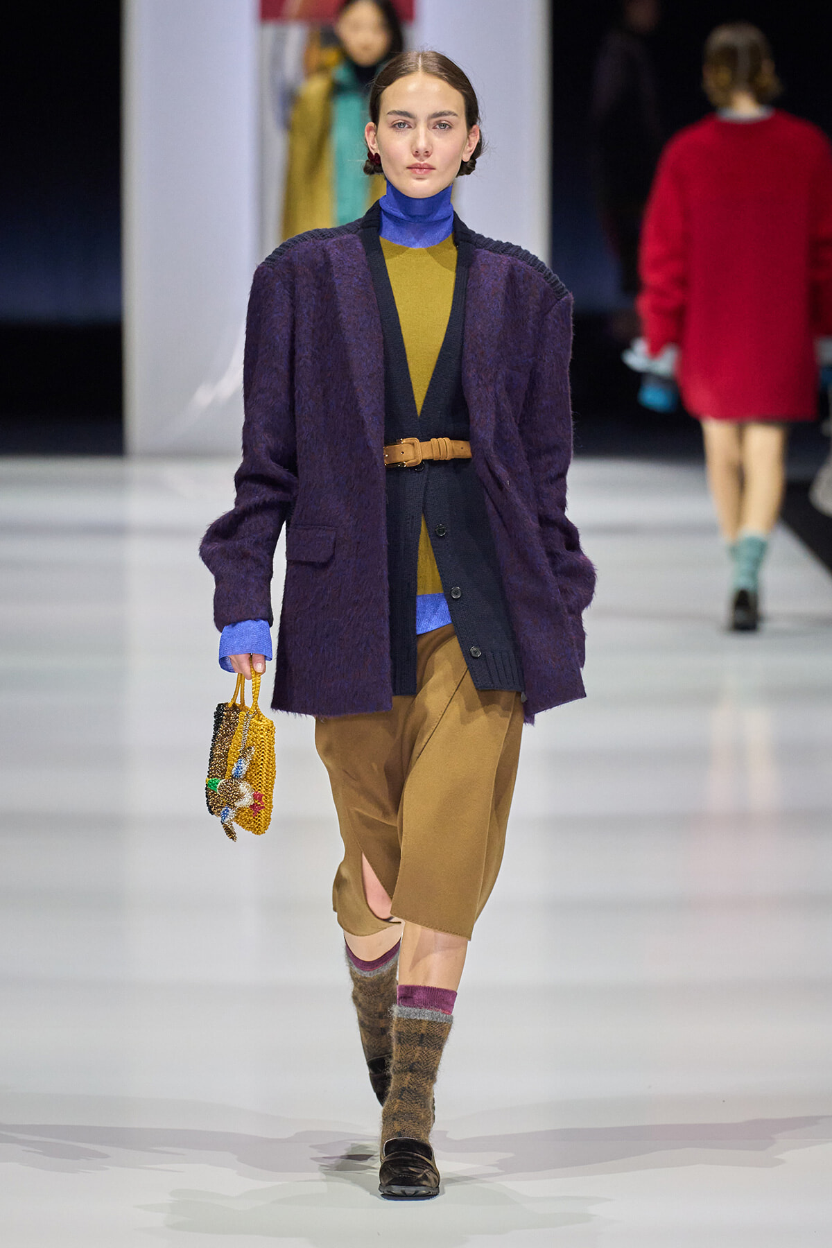 Model walking a fashion runway in a layered outfit: purple coat over mustard and blue knitwear, tan culottes, brown belt, beaded handbag.
