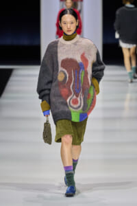 Model walks the runway in an oversized fuzzy sweater with abstract multicolor shapes, olive-green corduroy shorts, colorful socks, and blue shoes, carrying a beaded bag.