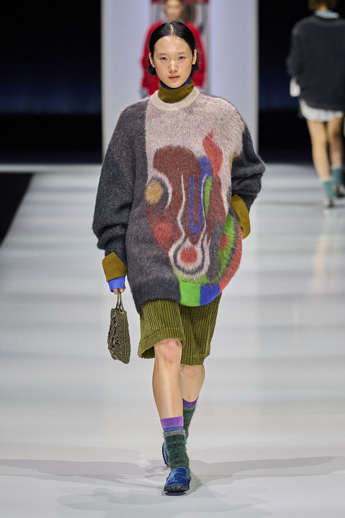 Model walks the runway in an oversized fuzzy sweater with abstract multicolor shapes, olive-green corduroy shorts, colorful socks, and blue shoes, carrying a beaded bag.