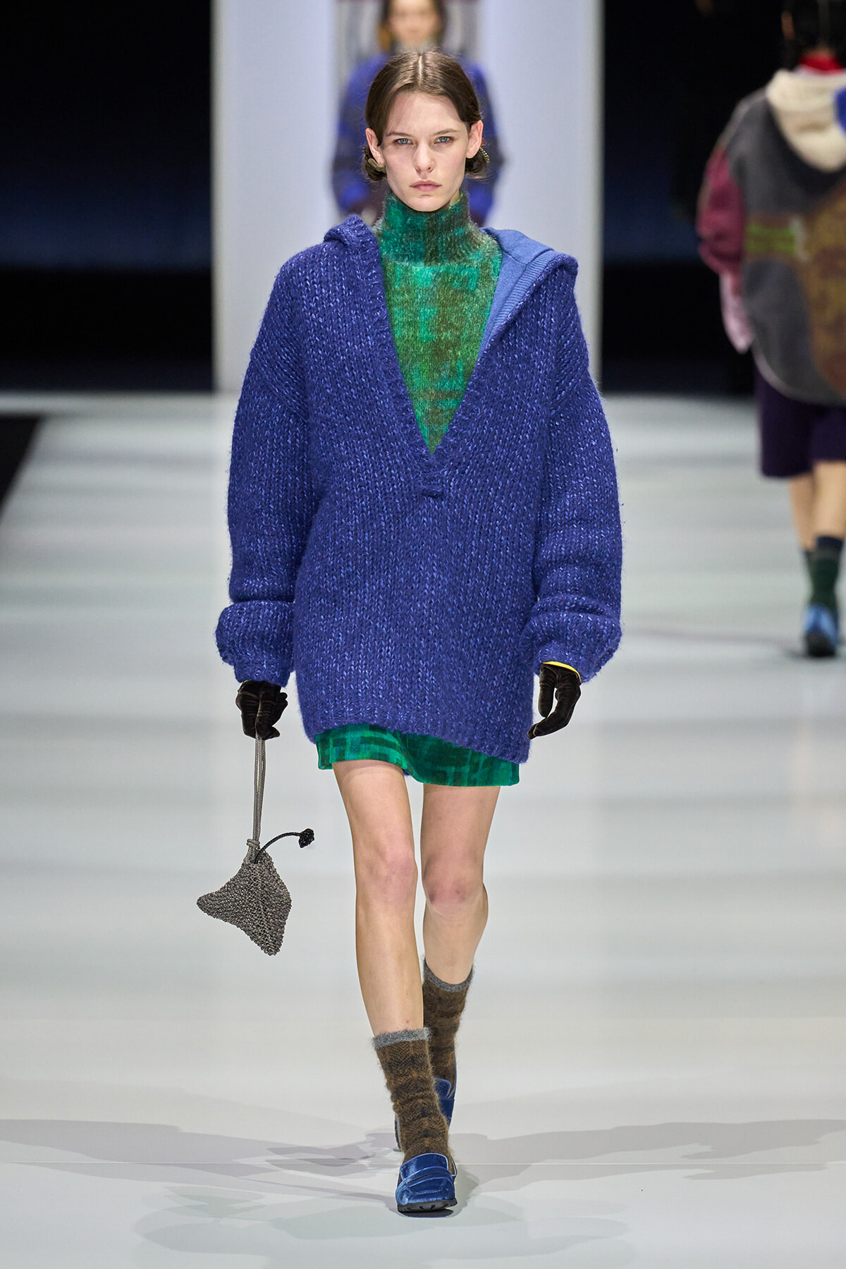 Model walks the runway in an oversized royal blue knit cardigan worn over a green plaid turtleneck dress, with brown fuzzy socks and blue loafers, carrying a small gray knit bag.