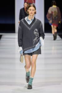 Runway model in a dark V-neck sweater over a light blue shirt, dark shorts, teal patterned socks, loafers, and a small gold beaded bag.