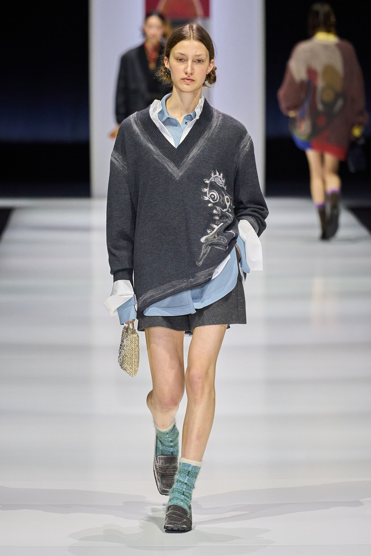 Runway model in a dark V-neck sweater over a light blue shirt, dark shorts, teal patterned socks, loafers, and a small gold beaded bag.