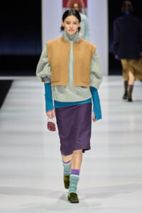 Model on a runway wearing a gray knit turtleneck with a camel sleeveless vest, a purple skirt, turquoise sleeves, and colorful socks with green velvet shoes, carrying a pink beaded purse.