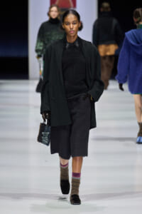 Model walks a fashion show wearing a dark oversized coat, black top, knee-length gray shorts, and fuzzy socks with loafers, carrying a small beaded bag.