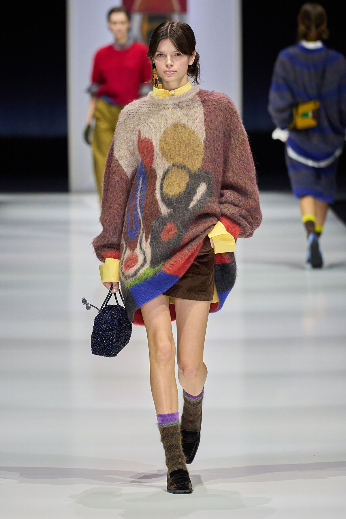 Model on a runway wearing a chunky abstract-pattern sweater, yellow-cuffed shirt, brown mini skirt, purple socks, brown boots, and a small navy handbag.