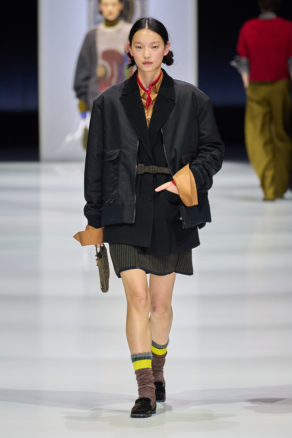 Model walking a runway in a black bomber jacket over a tan-cuffed outfit; wears a red neck scarf and belt, carrying a small beaded bag in her left hand.