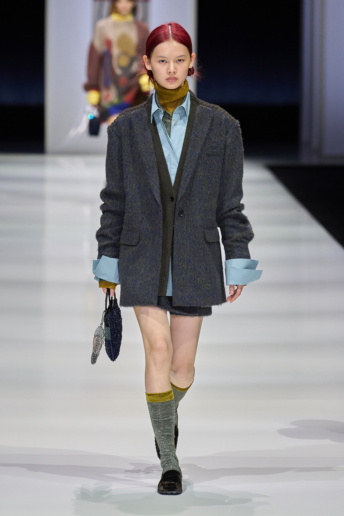 Model walks the runway in a oversized gray cardigan over a light blue shirt, mustard turtleneck peeking out, paired with matching blue cuffs, short shorts, and sparkly green socks with olive top accents while carrying a beaded handbag.