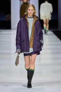 Model on a runway wearing an oversized purple jacket with tan lining over a light knit sweater and purple shorts, with striped knee-high socks and a beaded bag.