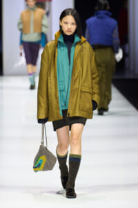 Model walking a runway in a teal inner jacket under a mustard oversized coat, carrying a knitted gray bag with a colorful design.