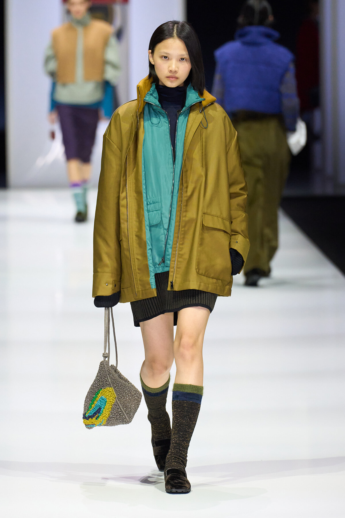 Model walking a runway in a teal inner jacket under a mustard oversized coat, carrying a knitted gray bag with a colorful design.
