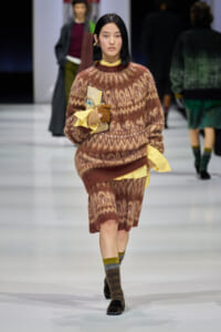 Model walking a fashion runway in a brown patterned fuzzy sweater and matching skirt with yellow accents, clutch in hand.