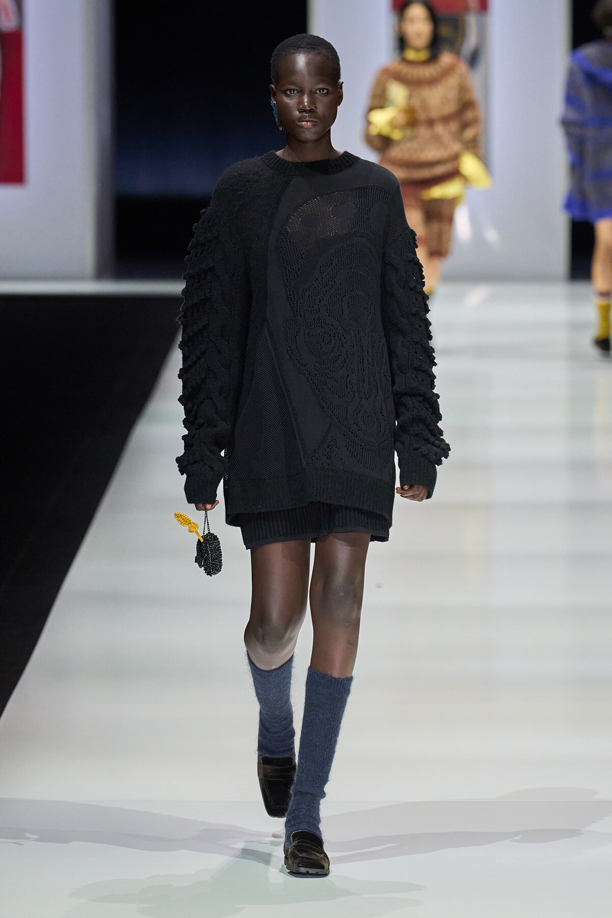 Model walking a fashion show runway in an oversized black knit sweater with textured sleeves, blue knee-high socks, brown loafers, and a small black beaded bag with a yellow tassel.