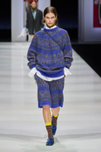 Model walks a fashion runway in a blue and brown patterned knit sweater and matching shorts, with a white shirt peeking at the hem and a diagonal crossbody strap.