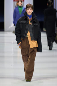 Model walking a fashion runway in a black cropped jacket over tan shirt and brown cargo pants, blue turtleneck visible at neck.
