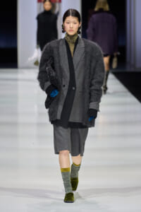Runway model in a large charcoal fuzzy cardigan over a gray suit with a matching knee-length skirt, layered with a black scarf and blue gloves, carrying a sparkly black clutch.