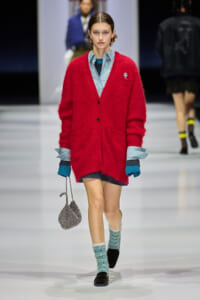 Model walking a runway in a bright red fuzzy cardigan over a light blue button-down shirt, with blue layered cuffs, mint socks, black loafers, and a glittery silver bag.