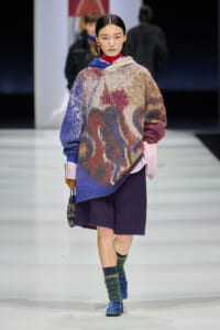 Model walking runway in a oversized multicolor fuzzy poncho-like sweater with purple shorts, red turtleneck, and bold earrings, carrying a small patterned bag.