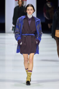 Runway model wearing a burgundy double-breasted coat cinched with a blue belt, over a darker purple shirt, with a bright blue knit cardigan layered over.