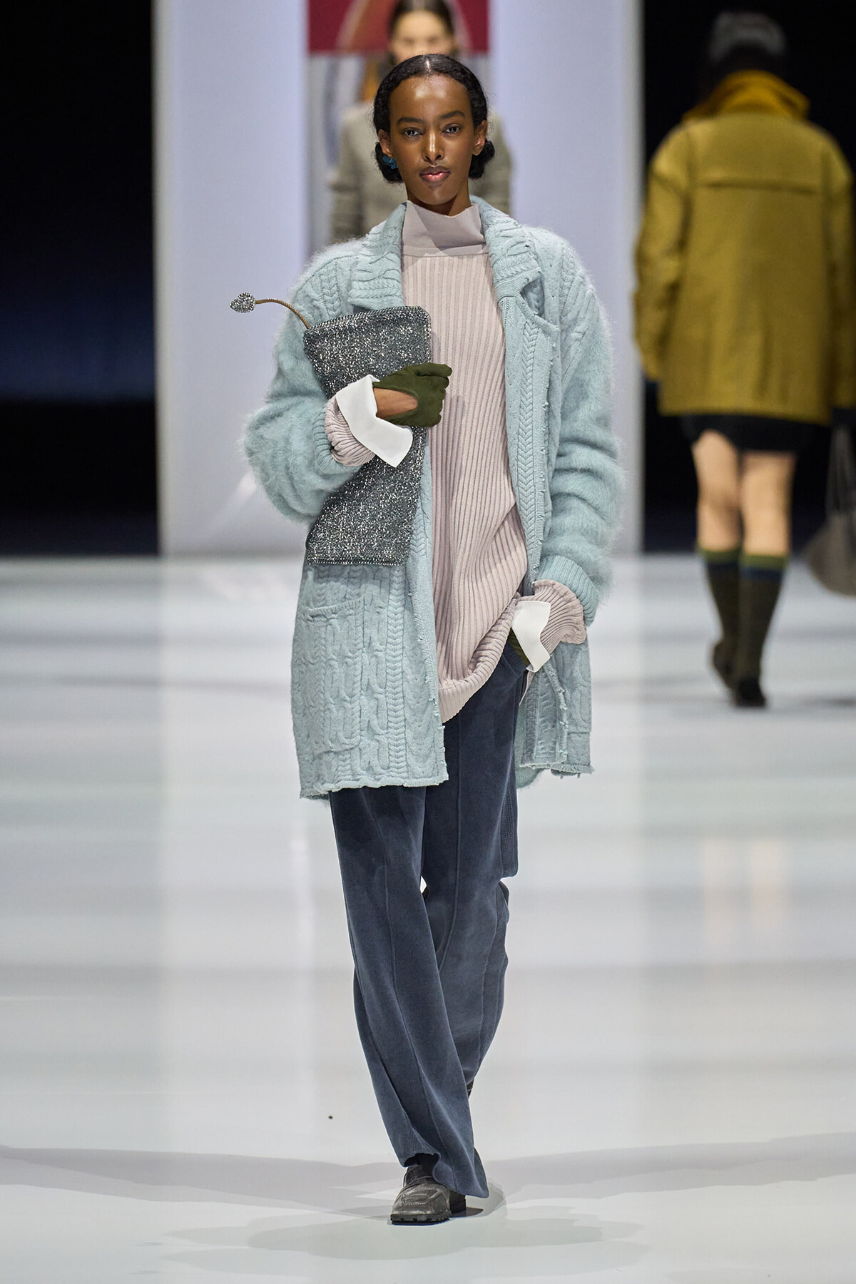 Model on a runway wearing a pale blue oversized knit coat over a beige turtleneck and blue wide-leg pants, clutch in hand.