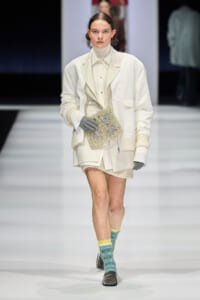 Model on a fashion runway in an oversized white coat and turtleneck, carrying a beaded beige handbag, gray gloves, and mint-green patterned socks with loafers.