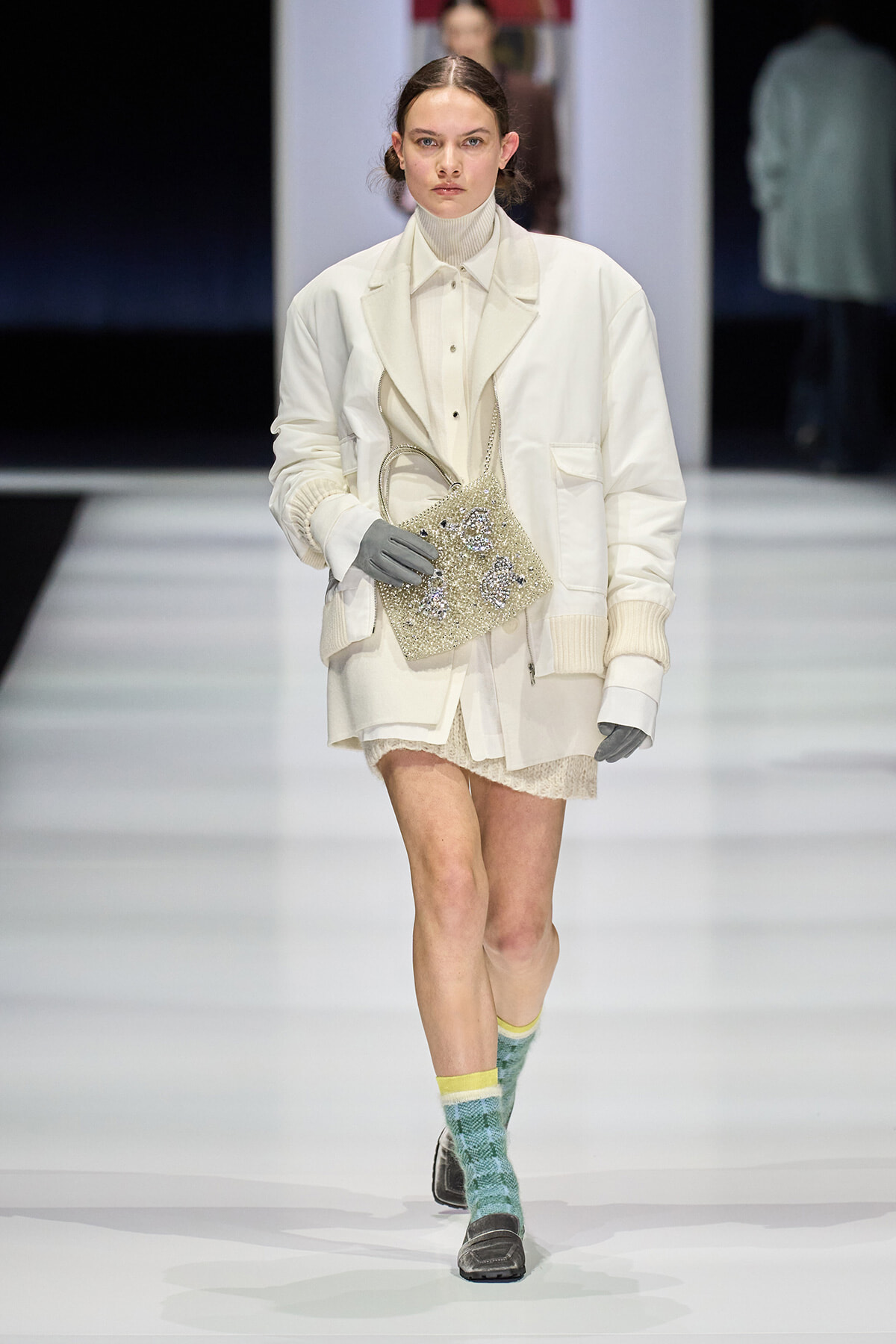 Model on a fashion runway in an oversized white coat and turtleneck, carrying a beaded beige handbag, gray gloves, and mint-green patterned socks with loafers.