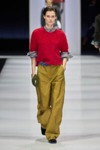 Model walking a runway in a bright red fuzzy knit sweater paired with olive wide-leg pants, blue belt, gray shirt sleeves peeking, and green glove on one hand