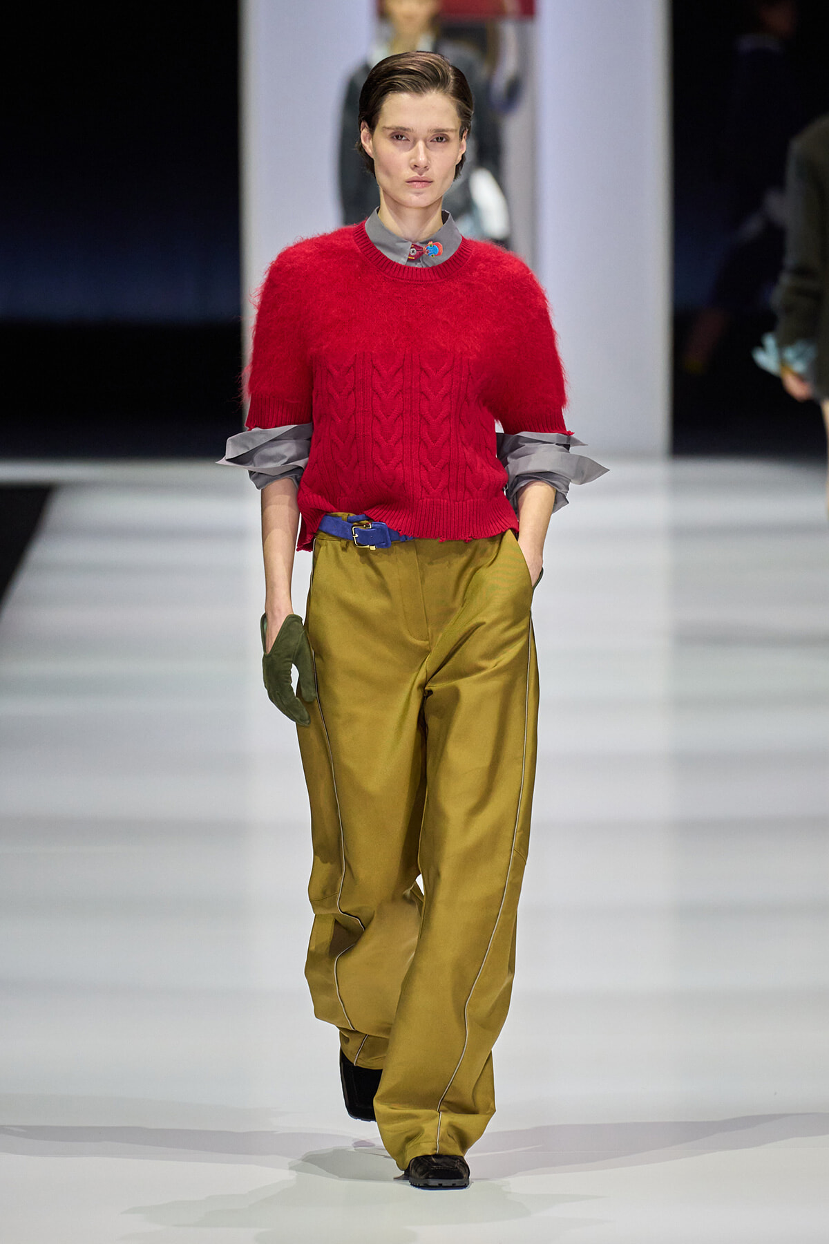 Model walking a runway in a bright red fuzzy knit sweater paired with olive wide-leg pants, blue belt, gray shirt sleeves peeking, and green glove on one hand