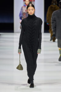 Model on a runway wearing a long black cable-knit turtleneck dress with metallic cuffs and a small beaded handbag.