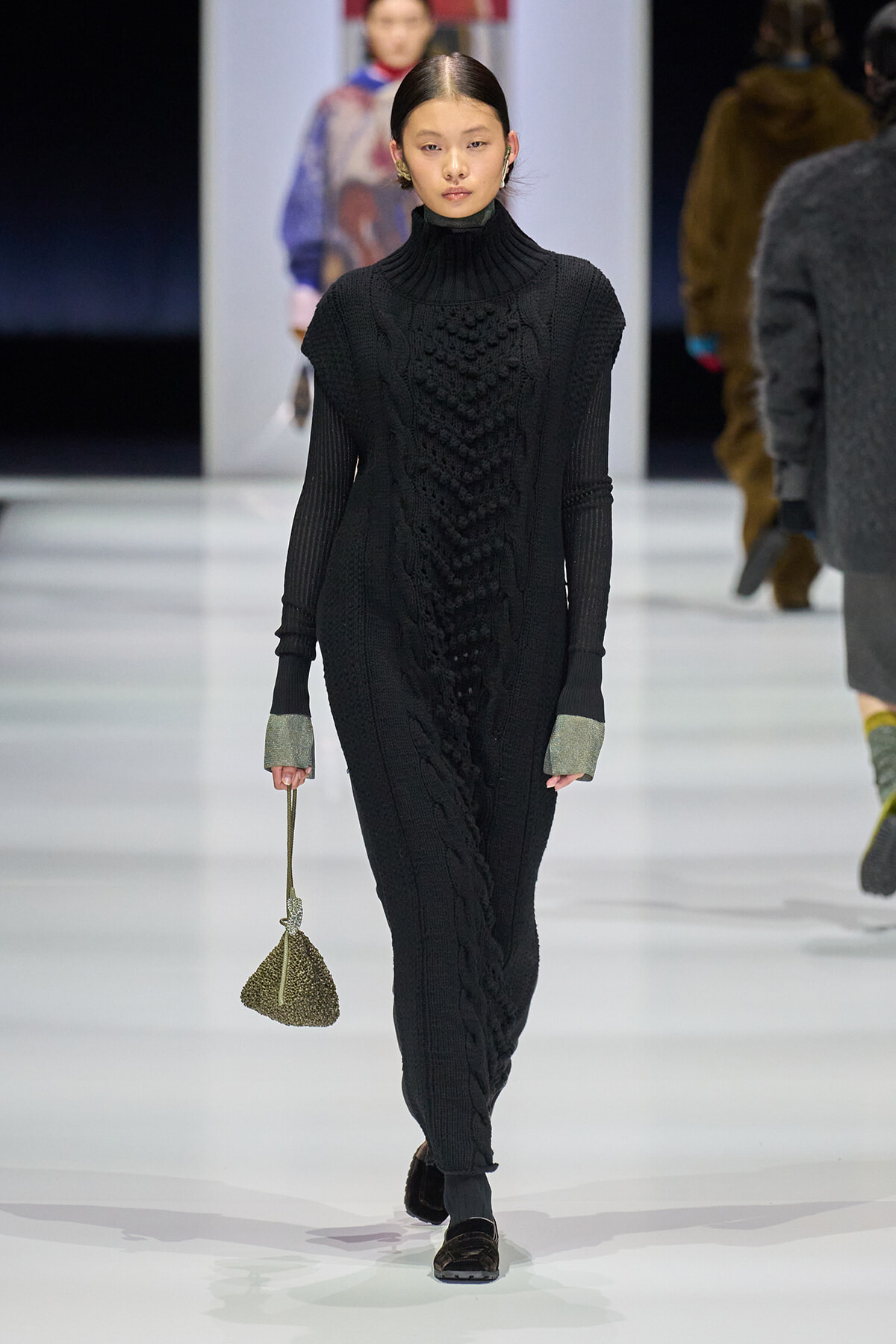 Model on a runway wearing a long black cable-knit turtleneck dress with metallic cuffs and a small beaded handbag.