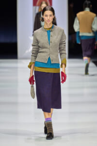 Model walks a fashion runway in a gray knit cardigan over a teal and mustard color-block top, paired with a dark purple midi skirt, red gloves, and brown shoes with multicolored socks; she carries a small beaded purse.