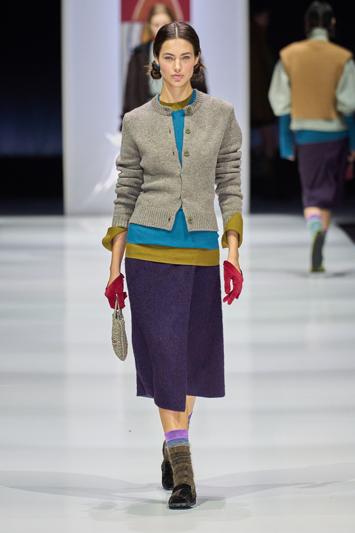 Model walks a fashion runway in a gray knit cardigan over a teal and mustard color-block top, paired with a dark purple midi skirt, red gloves, and brown shoes with multicolored socks; she carries a small beaded purse.