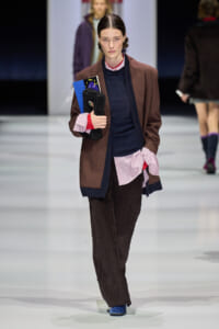 Model walking down a runway in a brown blazer over layered navy and pink shirts, holding a black clutch and a blue booklet.