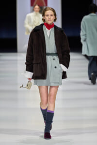 Model walks the runway in a pale blue belted dress under a dark brown corduroy coat, red turtleneck, and knee-high purple socks with burgundy loafers. She carries a small beige handbag.