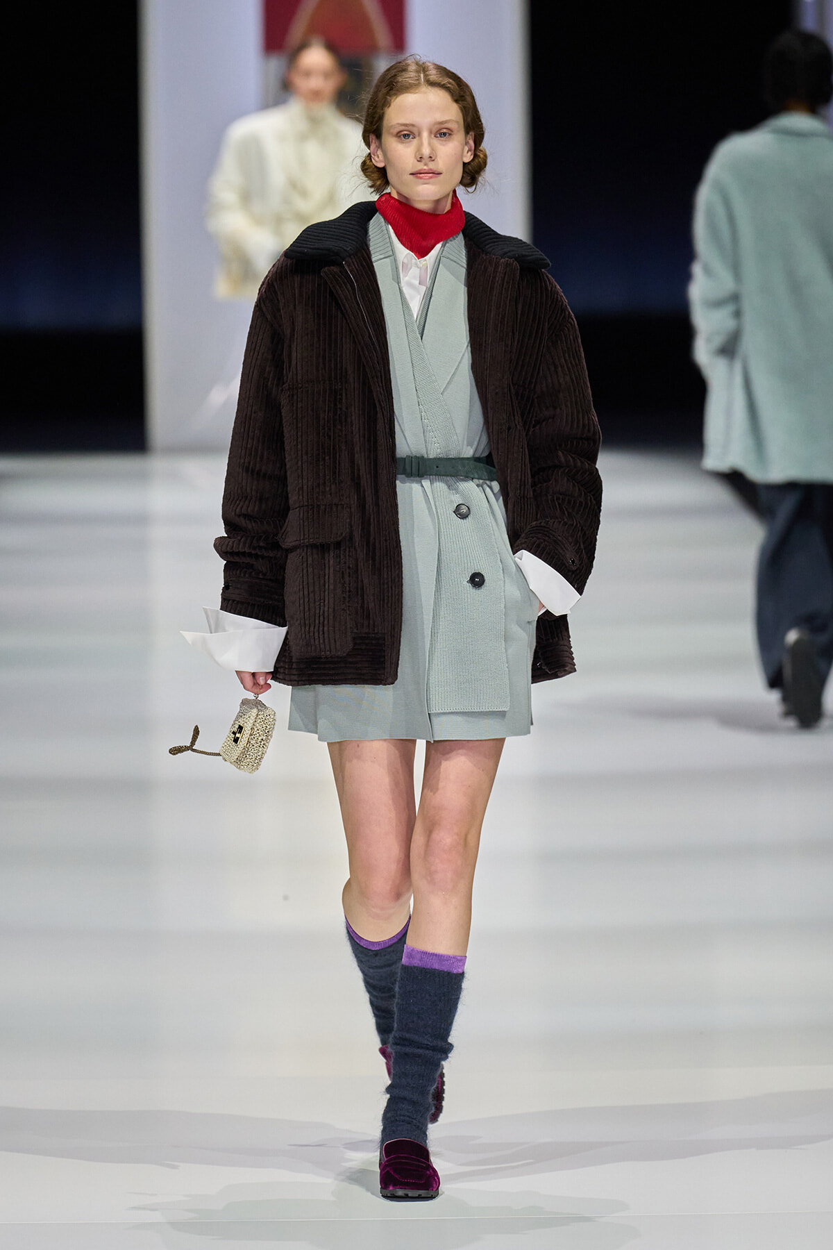 Model walks the runway in a pale blue belted dress under a dark brown corduroy coat, red turtleneck, and knee-high purple socks with burgundy loafers. She carries a small beige handbag.