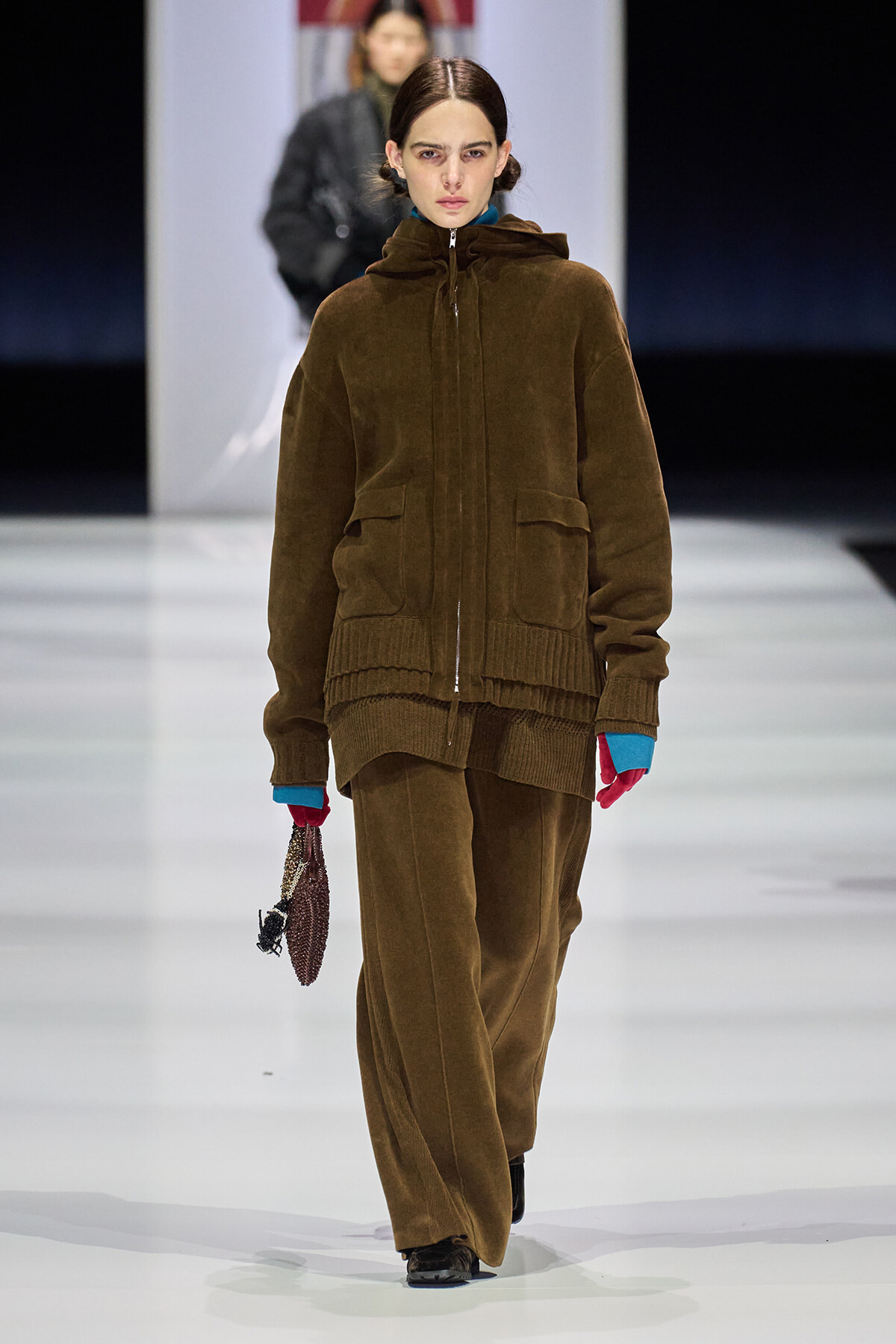 Model walking a fashion runway in a brown velour zip-hooded jacket and matching wide-leg pants, with bright blue cuffs peeking from sleeves and pink gloves, carrying a beaded purse.