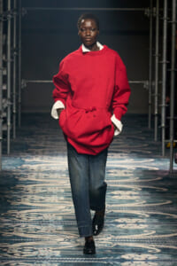 Model walking a runway in an oversized red knit sweater over a white shirt, blue jeans, and black loafers.