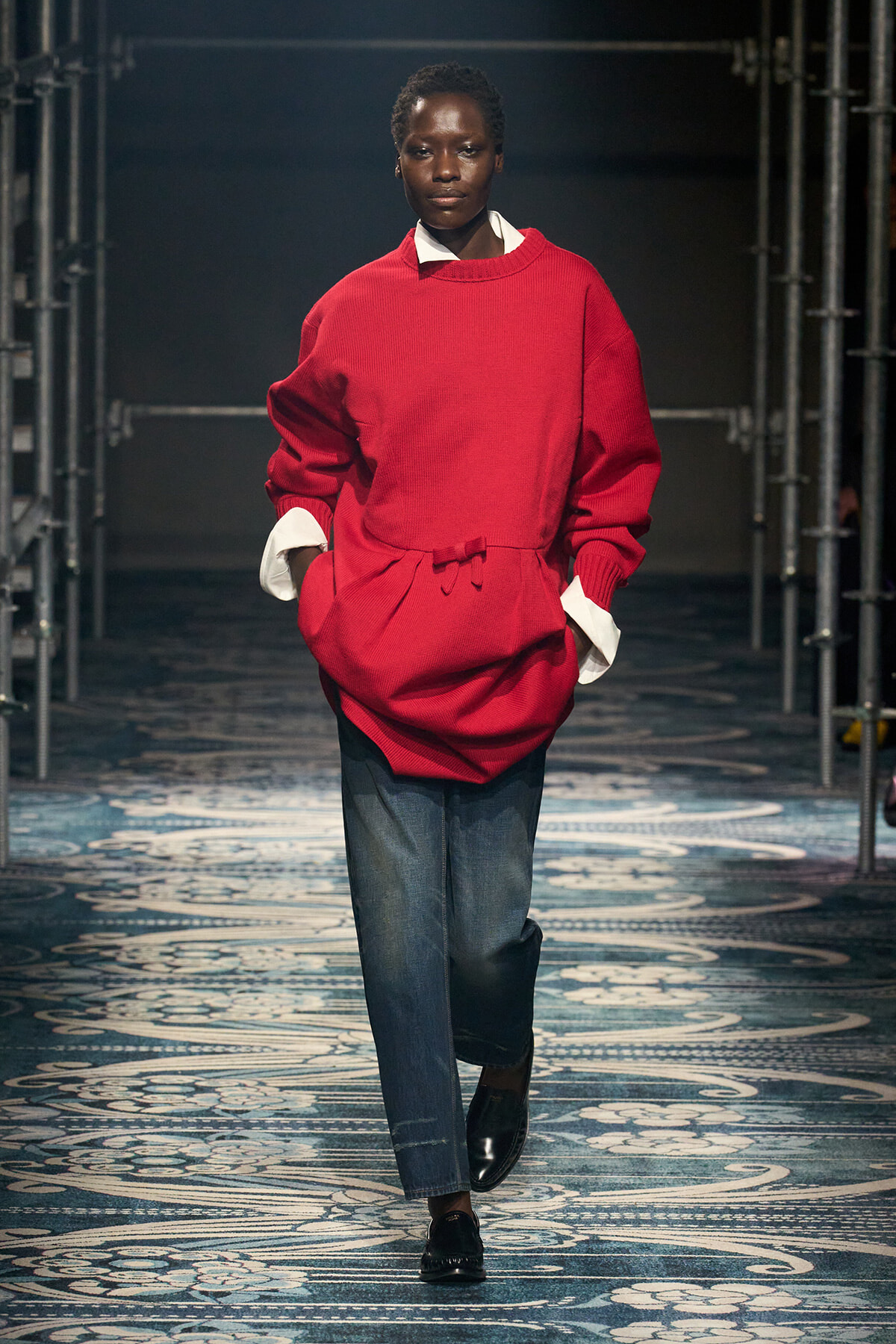 Model walking a runway in an oversized red knit sweater over a white shirt, blue jeans, and black loafers.