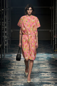 Model walking a fashion runway in a pink floral dress with a matching belt, carrying a small black handbag and wearing beige pointed heels.